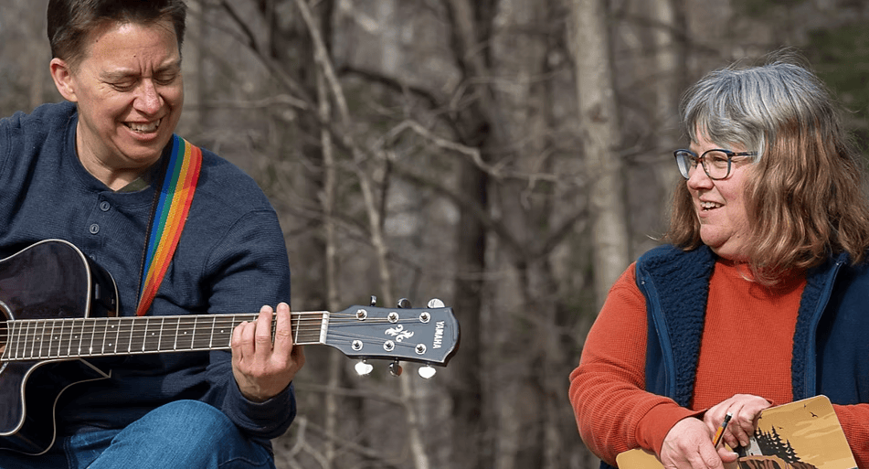 two people sit outdoors in front of bare trees; one plays a black acoustic guitar and the other holds a stringed instrument, both smiling as they interact.