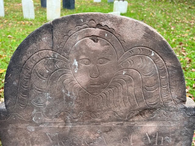 close up of an old gravestone featuring a carved face with wings or rays extending outward, set in a grassy cemetery with other blurred headstones in the background.