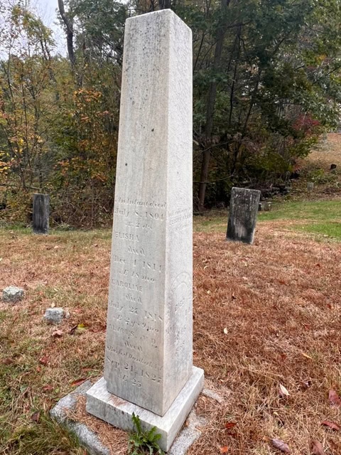tall, weathered stone obelisk gravestone stands in a grassy cemetery with trees and other headstones in the background.