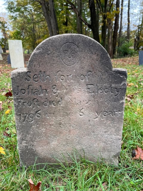 old gravestone in a grassy cemetery marking the grave of seth, son of josiah and electy, who died in march 1796 at age 6. other gravestones and trees are visible in the background.