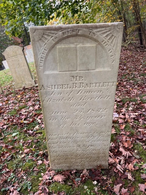 a weathered gravestone for ashel b. bartlett, who died june 25, 1832 at age 34, stands among fallen leaves in a wooded cemetery.