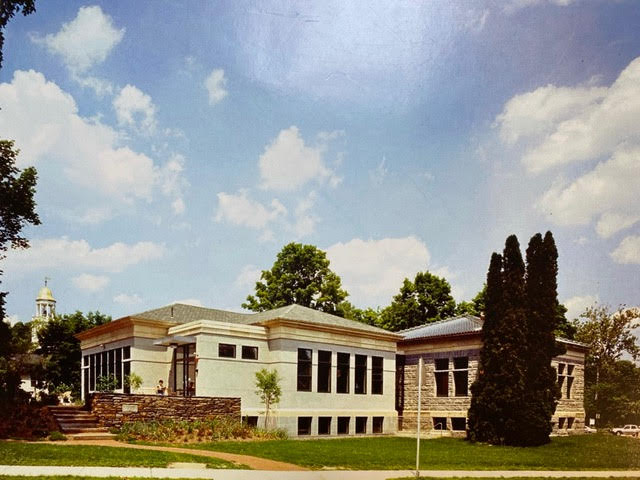 a one story stone and stucco building with large windows sits on a landscaped lawn under a partly cloudy sky. trees and a church steeple are visible in the background.