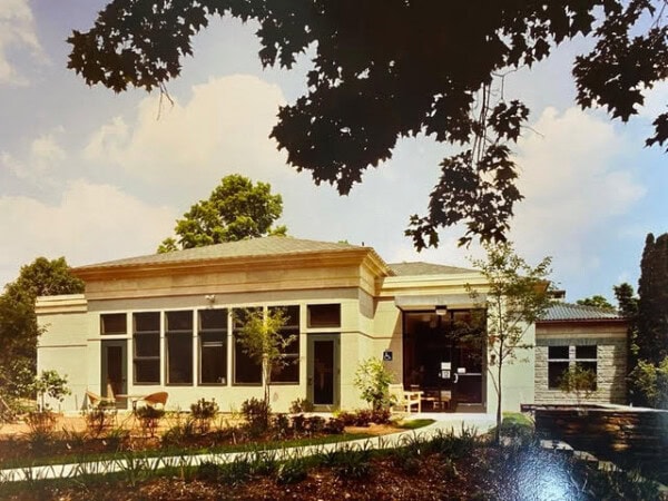 a single story building with large windows and a small entrance surrounded by landscaping and trees under a partly cloudy sky.