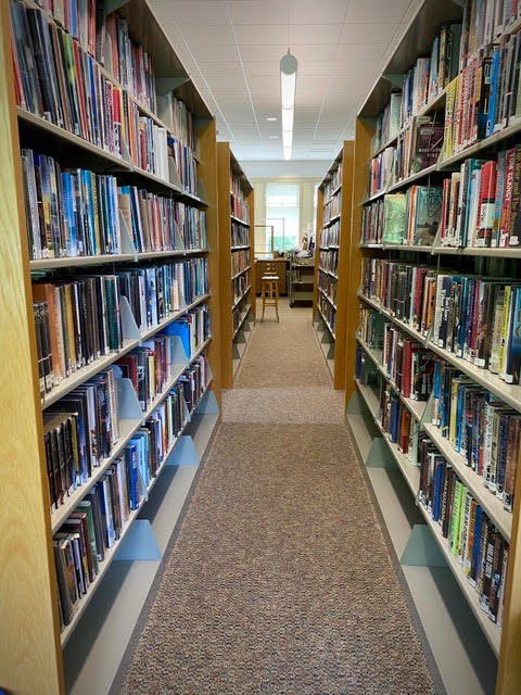 a carpeted aisle between tall bookshelves filled with books in a library, leading to a window with a table and chair at the end.