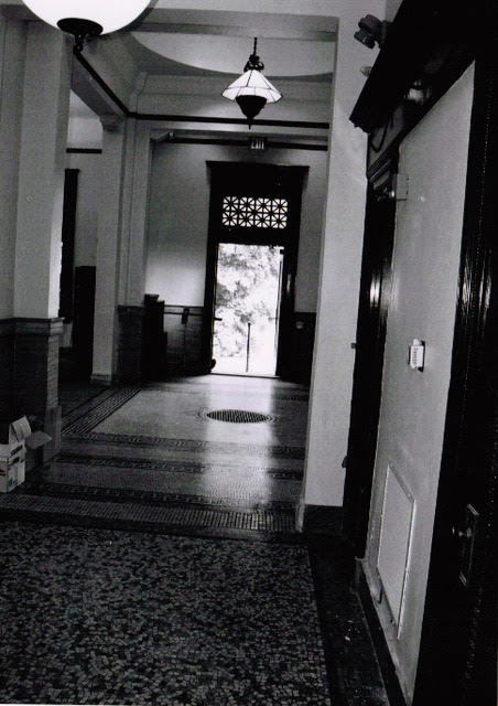 black and white photo of an empty hallway with tiled floors, high ceilings, hanging lights, and an open door at the far end letting in natural light.
