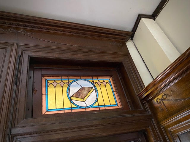 stained glass window with a central image of a book in a circular frame, surrounded by yellow and blue geometric designs, set in a wooden wall.