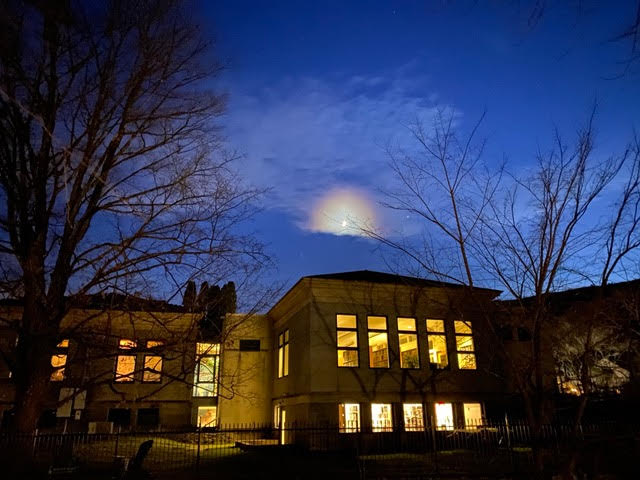 a building with illuminated windows at night, framed by leafless trees, under a dark blue sky with a glowing cloud.