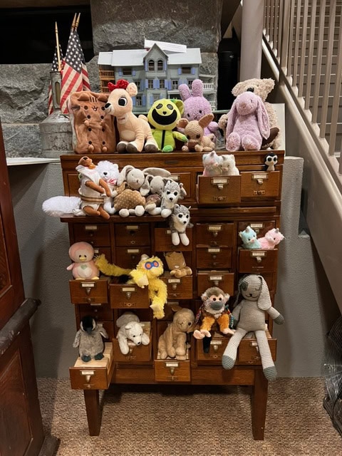 a wooden card catalog holds various stuffed animals and plush toys, with some sitting on top and others placed inside open drawers. a model house and two us flags are on top.