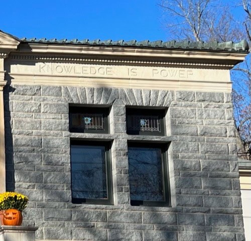 a stone building with two windows features the engraved phrase “knowledge is power” above them. a planter with yellow flowers and a carved pumpkin sits on a ledge below.