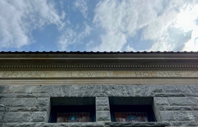 stone building facade with the names "bryant lowell holmes" engraved above two stained glass windows, set against a partly cloudy sky.