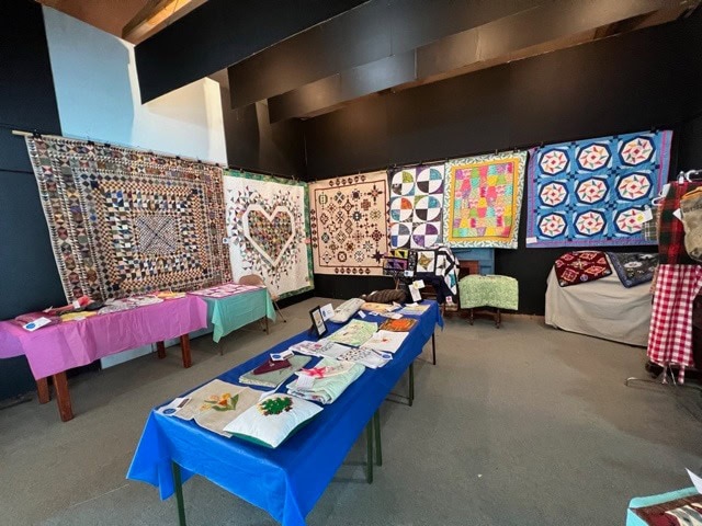 room displaying various colorful quilts and patchwork blankets hung on walls and draped over tables, with additional crafted textiles arranged on tabletops.