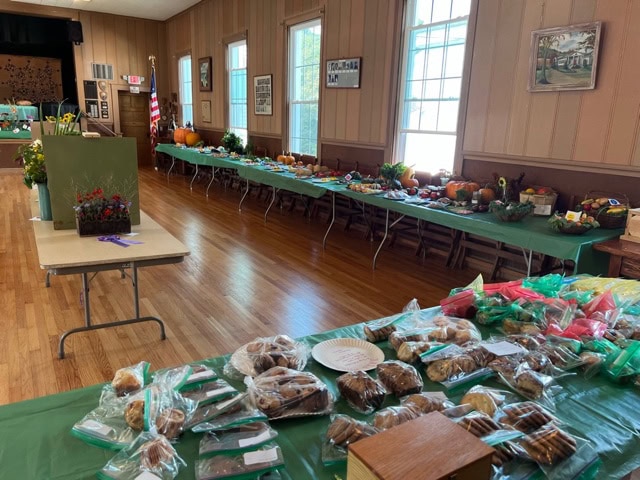 a community hall with long tables displaying baked goods, plants, and produce, all set up for what appears to be a local fair or market.