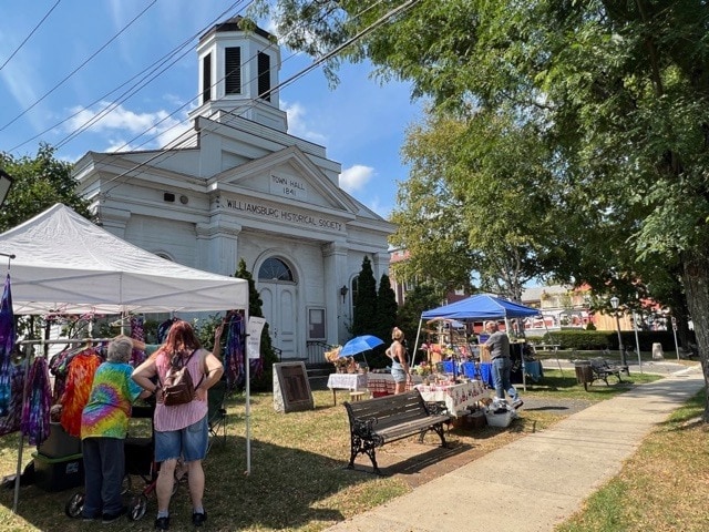 people browse outdoor vendor booths in front of the willimantic historical society building on a sunny day.