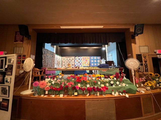 a stage displays colorful quilts in the background and jars of flowers with labels in the foreground, with two large fans on either side.