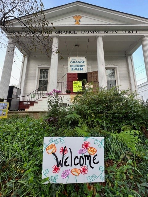 a white building labeled "williamsburg grange community hall" with a "grange community fair" banner and a colorful "welcome" sign in front, surrounded by plants.