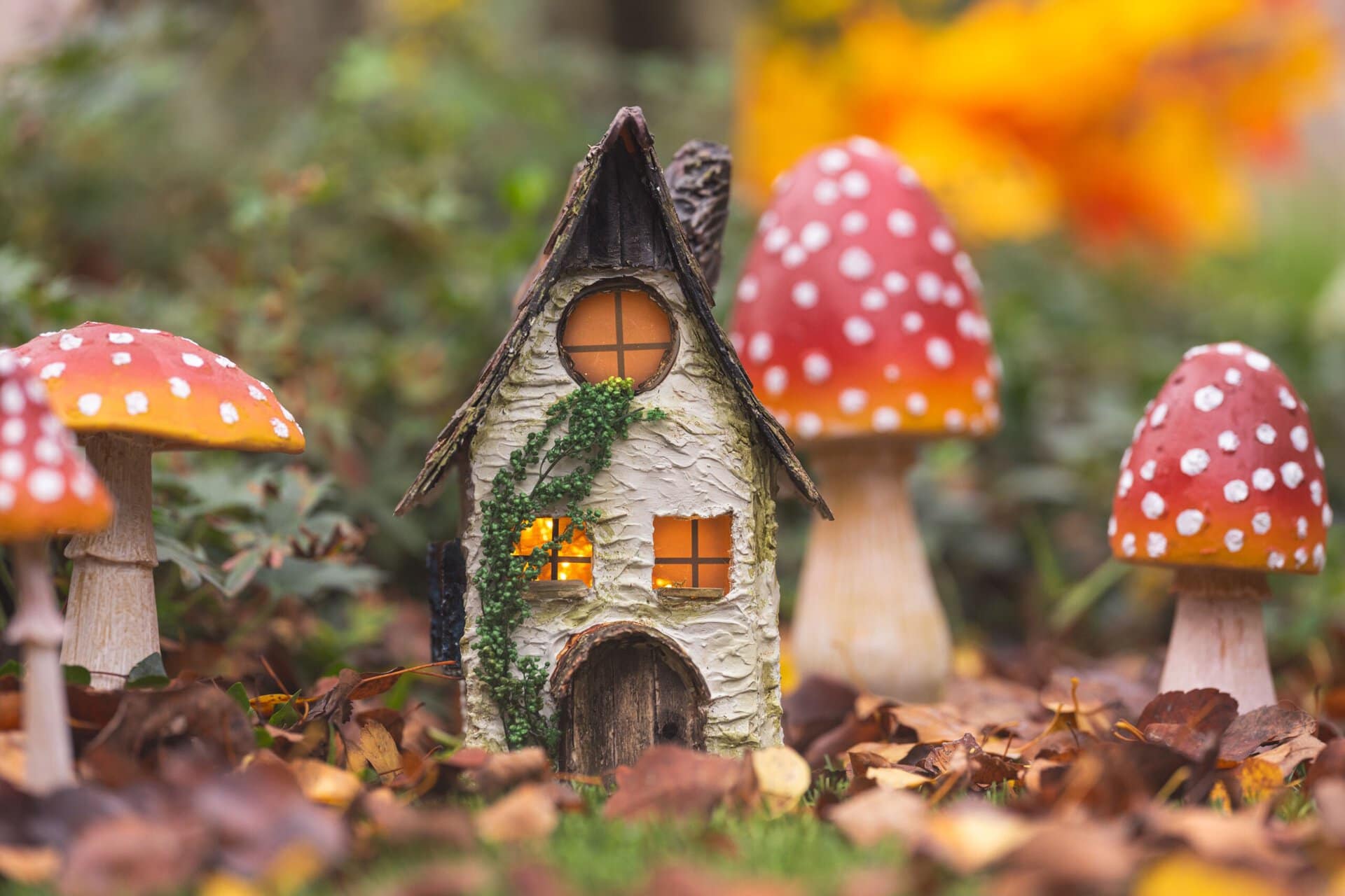 a miniature fairy house with lit windows surrounded by three decorative toadstool mushrooms in a bed of autumn leaves.