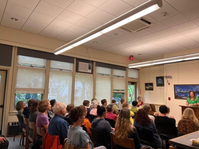 a group of people sit facing a speaker at the front of a well lit room with large windows and framed pictures on the walls.