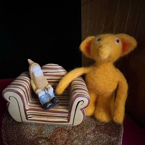 a felted yellow creature stands beside a striped miniature couch with a small gnome figurine seated on it.