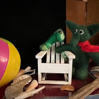 two green felt animal toys sit near a small white wooden chair, with a colorful beach ball and seashells scattered around them.