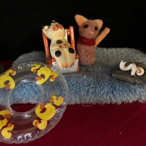 a small toy cat in a chair, a felted brown cat, a rubber ring with yellow ducks, and a curled white object on a gray base, all on a blue fuzzy mat with a red background.