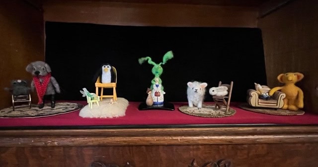 a display of six felt animal figurines on a shelf, each with small props such as chairs, rugs, and a couch, set against a black background.