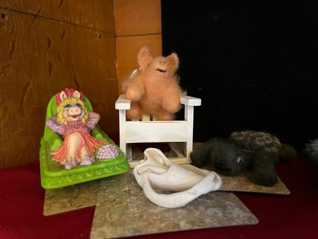 a small figurine of miss piggy on a green lounge chair sits next to wool animal figures and a small white chair on a red and brown surface with a wooden and black background.
