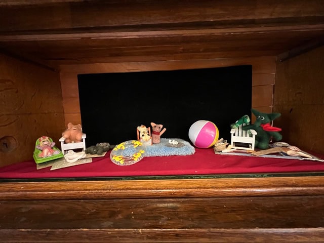a wooden shelf displays small animal figurines, a beach ball, miniature beds, shells, and a blue rug on a red surface with a black backdrop.