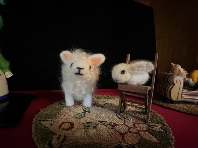 a small felted sheep stands on a rug next to a felted rabbit sitting on a miniature wooden chair, with a couch and other objects in the background.