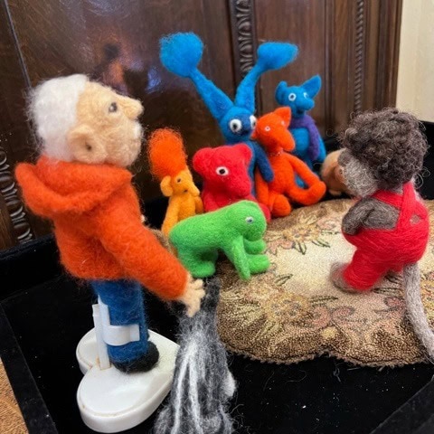 a felted wool figure of an older man faces colorful felted animal figures seated in a semicircle, while another felted person in red overalls sits nearby.