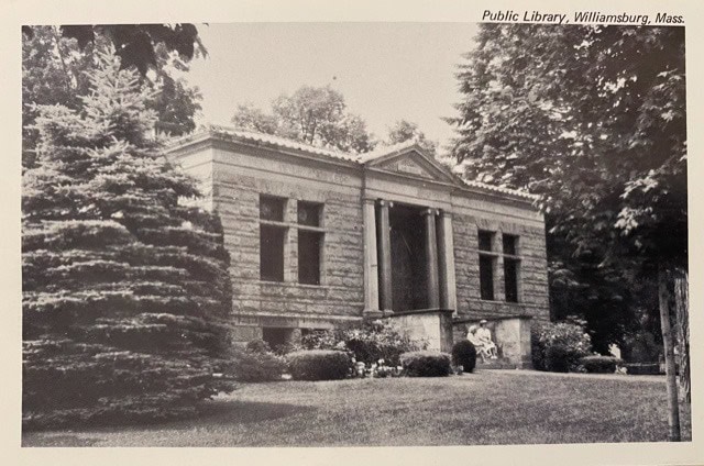 black and white photo of the williamsburg, mass. public library, a stone building surrounded by trees and shrubs, with a person seated near the entrance.