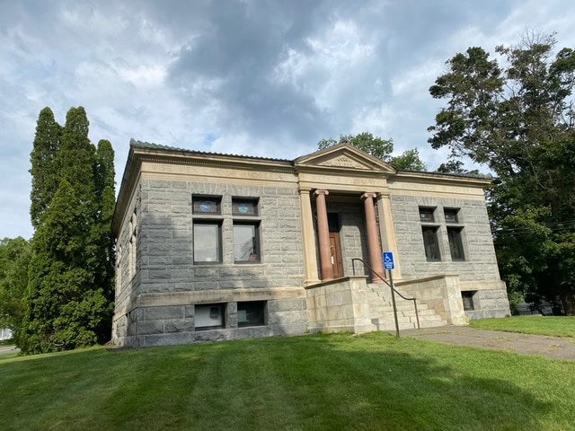 stone building with columns at the entrance, elevated by stairs, surrounded by grass and trees, under a cloudy sky with a visible accessible parking sign.