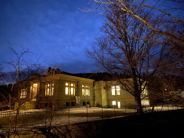 a building with illuminated windows sits behind bare trees and a fence at dusk, under a deep blue, cloudy sky.