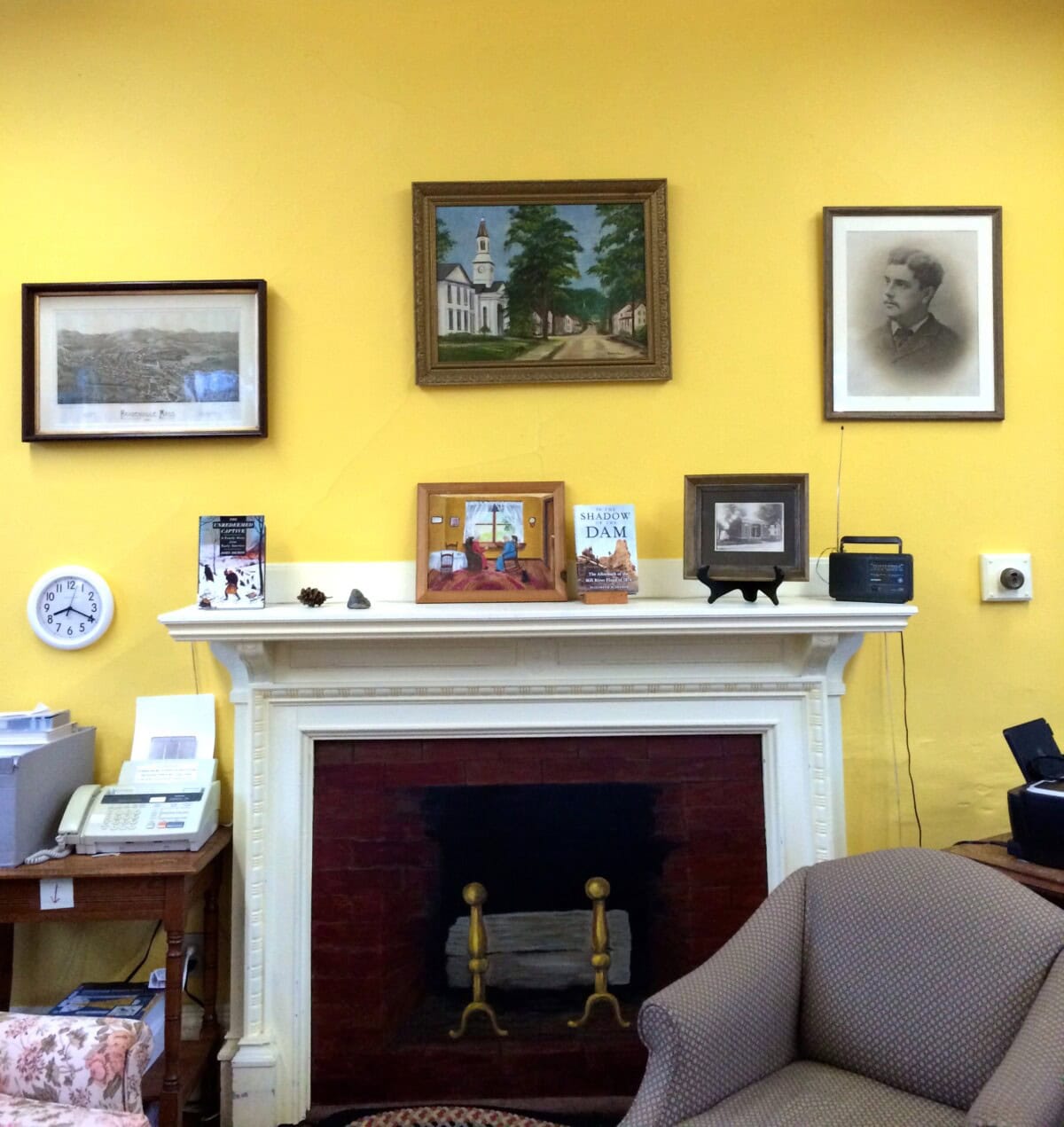 A yellow-walled room with a white fireplace, books, framed art and photos above the mantle, a radio, a clock, and an armchair in the foreground.
