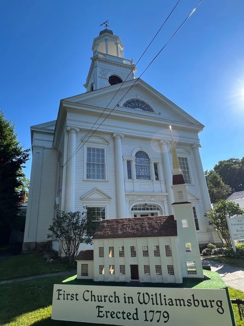 a white colonial style church with columns and a steeple; a sign in front reads "first church in williamsburg erected 1779" with a model of the church displayed.