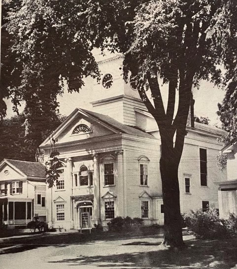 a large, white, colonial style church with a clock tower stands behind trees, with an adjacent smaller house visible to the left.