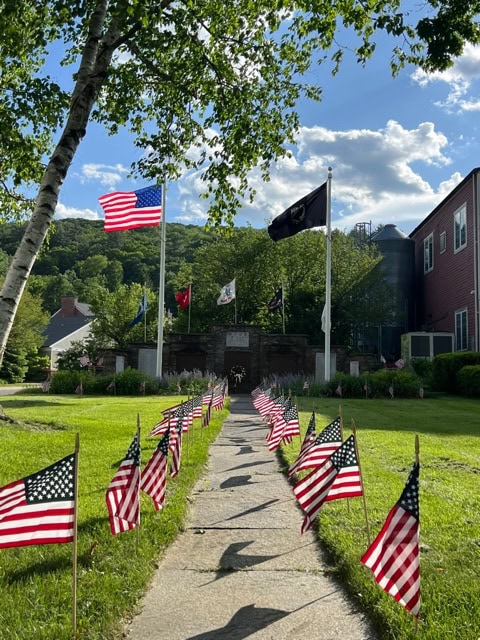 a walkway lined with small american flags leads to a memorial, with several flagpoles and buildings in the background on a sunny day.