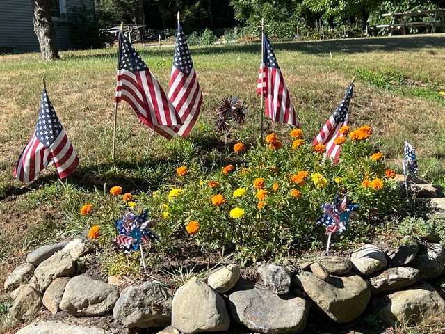 a small garden bed with yellow and orange flowers, surrounded by rocks, five american flags, and several star patterned pinwheels.