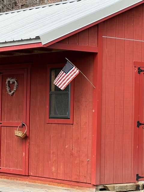a small american flag is mounted on the red exterior of a wooden building with a window, a door, a heart shaped wreath, and a hanging basket.