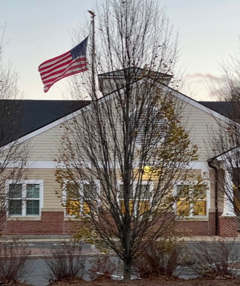 a u.s. flag flies at half staff in front of a beige building with white windows and a brick foundation, with leafless trees in the foreground.