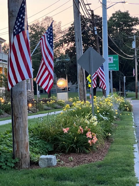 american flags hang from utility poles along a street with flower beds, green grass, and road signs visible in the background at dusk.