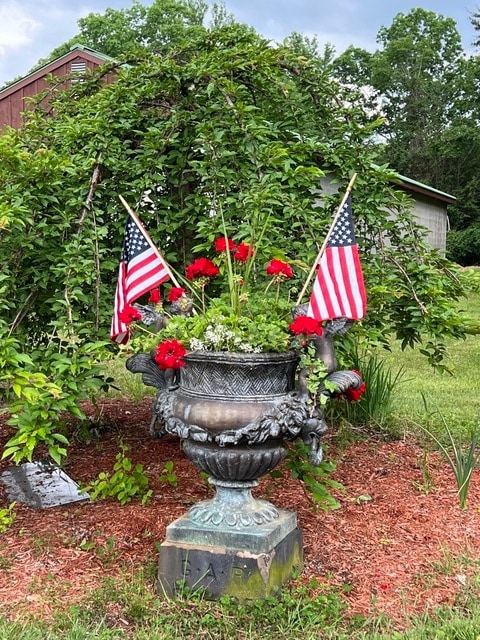 large decorative planter with red flowers and greenery, flanked by two small american flags, set in a mulched garden area with trees and buildings in the background.