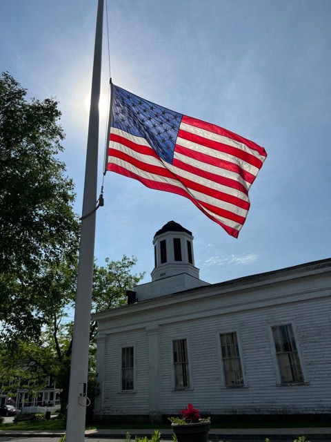 an american flag flies at half staff in front of a white historic building with a cupola, under a clear sky.