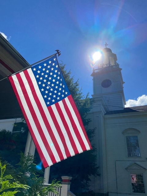 an american flag hangs outside a building with a tall clock tower, as sunlight shines brightly in the background.