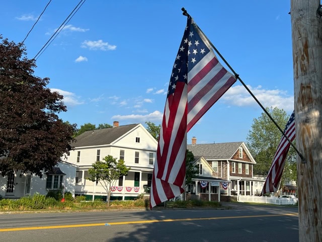 a street scene shows american flags and houses decorated with patriotic bunting on a sunny day with a blue sky.
