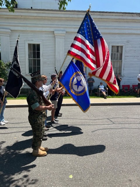 four people march in a parade holding the american flag, a blue military flag, and a black flag, with bystanders seated along the street in front of a white building.