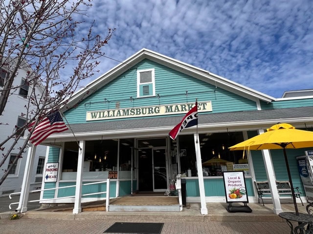 williamsburg market storefront with american flags, a sign advertising local produce, and an outdoor seating area with a yellow umbrella under a partly cloudy sky.