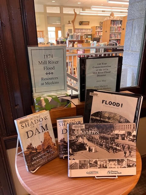 a library display features books and resources about the 1874 mill river flood, with a sign highlighting its 150 year commemoration and related materials available at meckins.