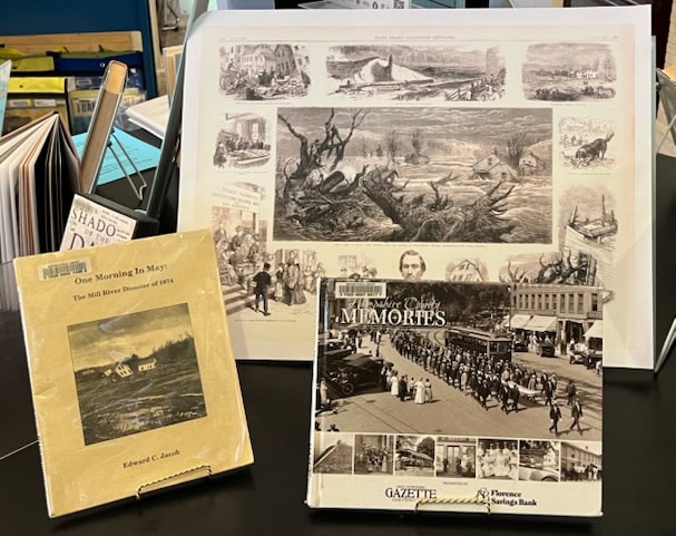three historical books and prints are displayed upright on stands, featuring black and white images and illustrations of past events and scenes.