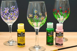 three wine glasses decorated with hand painted flowers stand behind bottles of yellow, green, and pink acrylic paint and a paintbrush on a wooden surface.