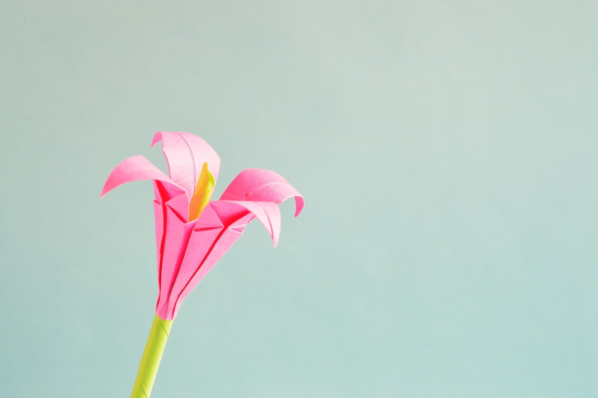 a pink origami flower with a yellow center on a green stem, set against a plain light blue background.